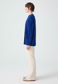 Kvinna i profil som bär en blå knäpp cardigan, off-white byxor och beige sneakers, stående mot en enkel ljus bakgrund.
