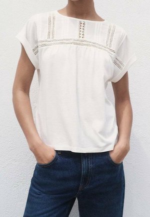 Biały top z krótkim rękawem i dekoltem w łódkę, ozdobiony teksturowaną lamówką oraz koronkowymi detalami na wysokości klatki piersiowej, noszony z ciemnoniebieskimi dżinsami.