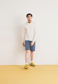 Helle beige Langarmbluse, blaue Shorts und gelbe Sportschuhe. Steht auf einem gelben Boden vor einer weißen Wand.