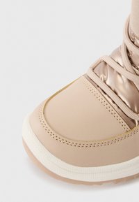 Beige waterdichte laars met een glanzende roségouden bovenkant, textuurlich lichaam, crème rubberen zool en bijpassende veters, met gestikte details.