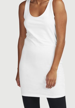 Kvinde iført en hvid ærmeløs tankkjole, der står mod en ensfarvet lys baggrund, med sorte leggings synlige under kjolen.