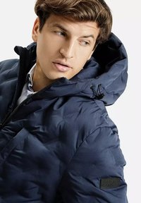Granatowa kurtka puchowa z kapturem, z zamkiem błyskawicznym i teksturowanym wypełnieniem. Zawiera naszywkę z logo na lewej rękawie.