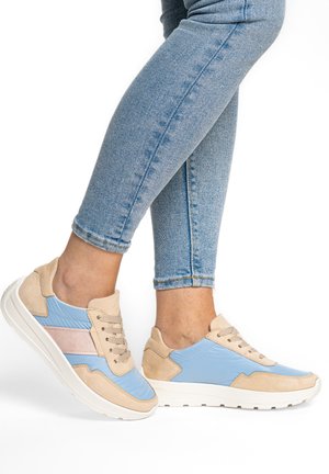 Person, die hellblaue und beige Sneaker mit rosa Streifen trägt, kombiniert mit hellblauen, eng anliegenden Jeans, stehend vor weißem Hintergrund.