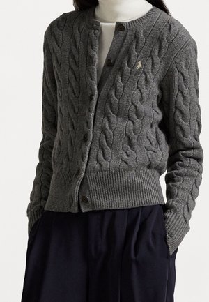 Mujer con un cárdigan de punto grueso gris abotonado sobre un suéter de cuello alto blanco y pantalones oscuros plisados, con las manos en los bolsillos.