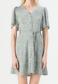 Femme portant une robe verte à manches courtes avec des pois blancs, boutonnée sur le devant et une ceinture en tissu nouée à la taille.