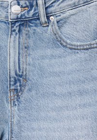 Jean en denim bleu clair en gros plan montrant la poche avant, un bouton en métal marqué "PULL&BEAR" et des détails de coutures orange.