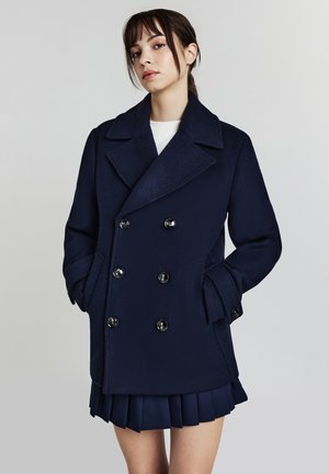 Cappotto peacoat in lana blu navy con chiusura a doppio petto, ampi revers e tasche laterali. Indossato sopra una mini gonna plissettata blu navy.