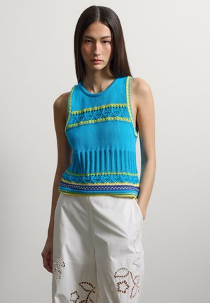 Femme portant un top en maille bleu sans manches avec des motifs jaunes et verts, associé à un pantalon blanc avec des découpes florales.