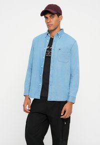 Ανοιχτό μπλε τζιν πουκάμισο με κουμπιά, μακριά μανίκια, τσέπη στο στήθος και μαύρη μπλούζα με γραφικά από κάτω. Το μοντέλο φοράει ένα μπορντό καπέλο. Μαύρο παντελόνι.