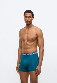 Ανδρικά μπόξερ σε τιρκουάζ από απαλό ύφασμα, με μπλε ζώνη μέσης που φέρει το λογότυπο "BJÖRN BORG". Σχεδιασμένα με εφαρμοστή γραμμή και λεία υφή.