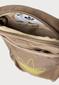 Καφέ υφασμάτινη τσάντα με υφή, που διαθέτει κίτρινο λογότυπο Adidas, άνοιγμα με φερμουάρ και λευκή εσωτερική ετικέτα με λογότυπο.