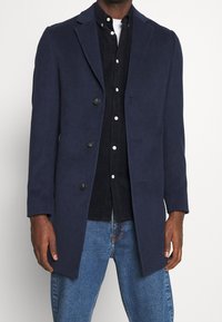 Cappotto in misto lana blu navy con design sartoriale, rever sciallati, chiusura con cinque bottoni e maniche a taglio dritto, indossato sopra una camicia scura e jeans.