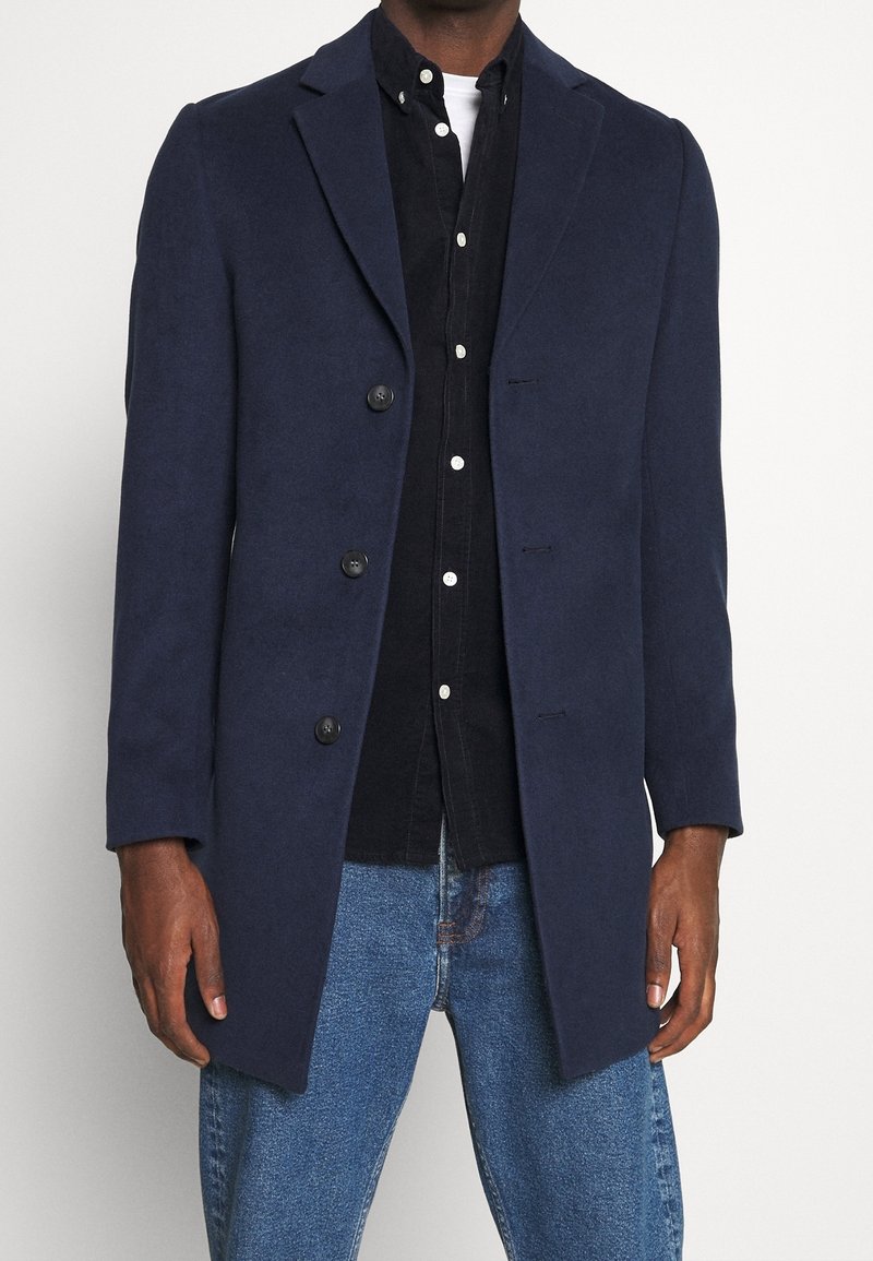 Cappotto in misto lana blu navy con design sartoriale, rever sciallati, chiusura con cinque bottoni e maniche a taglio dritto, indossato sopra una camicia scura e jeans.