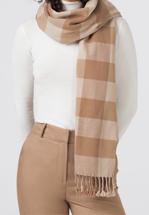 Femme portant une écharpe à carreaux beige et crème sur un col roulé blanc et un pantalon beige, l'écharpe pend librement avec des extrémités à franges.