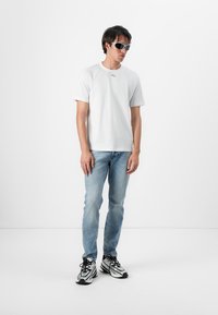 Tricou alb din bumbac cu un mic logo pe piept, blugi de denim bleumarin deschis și pantofi sport alb-negru. Model purtând ochelari de soare.