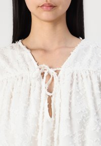 Weißes Blusenshirt aus strukturiertem Stoff mit fransigen Akzenten, einem Schlüsselloch-Ausschnitt mit einer Schleife und einem locker sitzenden, gerafften Design.