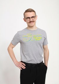 Grå bomulds t-shirt med gul og blå tekst "JT Racing USA" på forsiden, parret med sorte bukser og et sort bælte.