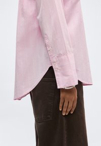 Άτομο που φοράει ροζ ανοιχτό πουκάμισο με μακριά μανίκια και κουμπωτά μανσέτα και σκούρο καφέ παντελόνι, με το χέρι χαλαρό δίπλα στο σώμα.