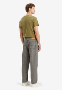 Camiseta de manga corta en verde oliva combinada con unos pantalones vaqueros grises de corte amplio. Presenta una tela texturizada y detalles en el bolsillo trasero. Zapatillas blancas.