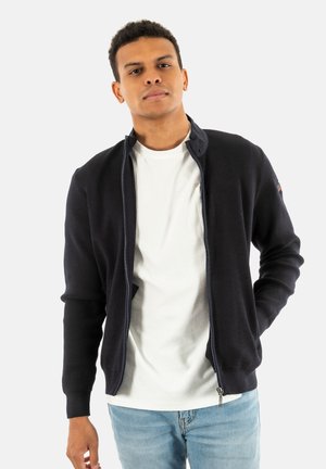 Veste zippée tricotée marine avec une texture, conçue avec un col montant, associée à un t-shirt blanc et un jean bleu clair.