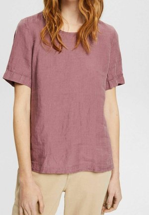 Frau trägt eine kurzärmlige Leinenbluse in staubrosa und beigefarbene Hosen, steht vor einem einfarbigen hellen Hintergrund.