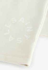 Šviesiai bežinė juostelėmis medžiaga, pasižyminti toniniu apvaliu logotipu su tekstu "SAINT LAURENT". Įtraukta "LONDON" šalia apvado.