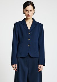 Marineblauer, maßgeschneiderter Blazer mit Reverskragen, fünf goldenen Knöpfen und zwei Vordertaschen. Weicher Stoff mit strukturierter Passform.