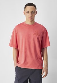 Camiseta rosa de gran tamaño y manga corta, hecha de algodón, con cuello redondo y un logo impreso que dice "EDUCACIÓN FÍSICA" en texto oscuro.