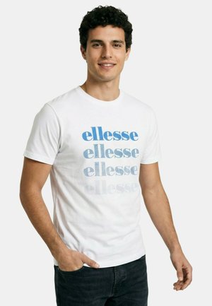 Homme souriant aux cheveux bouclés foncés portant un t-shirt blanc avec "ellesse" imprimé quatre fois en nuances de bleu, associé à un jean noir.