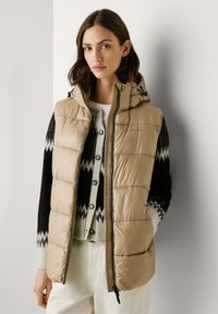 Beige puffer vest met een capuchon, met een geborduurd ontwerp, een rits aan de voorkant en een trekkoord. G gedragen over een zwarte en crèmekleurige gebloemde trui.