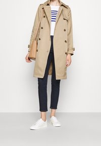 Osoba v béžovém trenčkotě, pruhovaném bílém a modrém tričku, tmavých slim kalhotách, bílých teniskách a s hnědou kabelkou přes rameno.