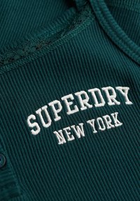 Top de tirantes en color verde azulado, con borde de encaje, que presenta bordado "SUPERDRY NEW YORK" en blanco. Incluye dos botones en el escote.