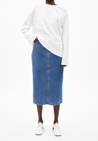 Persona che indossa una camicia bianca a maniche lunghe con la scritta "ARMEDANGELS", una gonna midi in denim blu, scarpe bianche con lacci, in piedi con una mano in tasca.