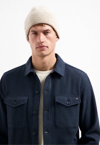 Marineblauwe button-up jas van gestructureerde stof met twee borstzakken, gecombineerd met een crèmekleurige gebreide muts. De jas heeft een kraag en knoopsluitingen.