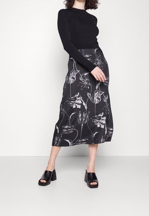 Kvinde iført en sort langærmet top, sort blomstret midinederdel og sorte åbne blockhælssko, stående på en lys gulv.