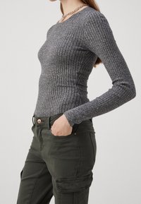Haut gris à manches longues et côtes, associé à un pantalon cargo vert olive avec poches avant et fermeture par bouton. Détail de collier en chaîne argentée.