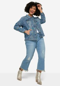 Veste en denim avec un motif de éclaboussures multicolores, fermeture à boutons, poches avant, associée à un jean bleu clair déchiré et des bottines métalliques.