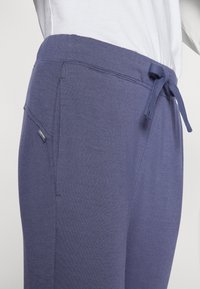 Lila Jogginghosen aus weichem Stoff mit elastischem Bund, Kordelzug und seitlicher Tasche. Glatte Textur mit minimalem Branding.
