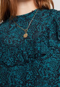 Žena v tyrkysovém a černém topu s abstraktním vzorem a volánkovými detaily, doplněná zlatým náhrdelníkem s kulatým přívěskem.