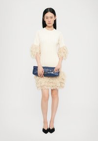 Robe en tricot côtelé crème avec des manches à franges et une jupe à volants frangés, associée à une pochette texturée bleu foncé et des chaussures noires à bout pointu.