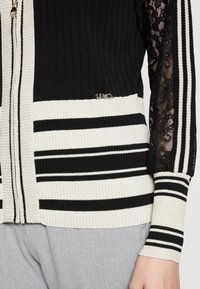 Zwart-wit gestreepte gebreide vest met geribbelde textuur, mouwen van kant en een kleine metalen logospeld op de taille, gedragen met grijze broek.