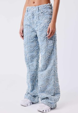 Jeans azzurri chiari a gamba larga con motivi floreali testurizzati, indossati con sneakers bianche chunky, mostrati dalla vita ai piedi.