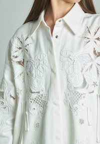 Camisa blanca bordada, con patrones florales, adornos texturizados, cuello con solapa y botones a presión visibles.