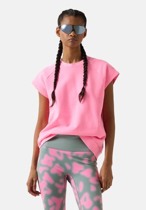 Frau mit geflochtenem Haar, die reflektierende Sonnenbrillen trägt, ein locker sitzendes, pinkes ärmelloses Oberteil und gemusterte pink-graue Leggings.