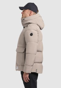 Puffer jacket in light beige with a hood, featuring a black logo patch, zippered front pocket, and elastic cuffs. Smooth, padded texture.