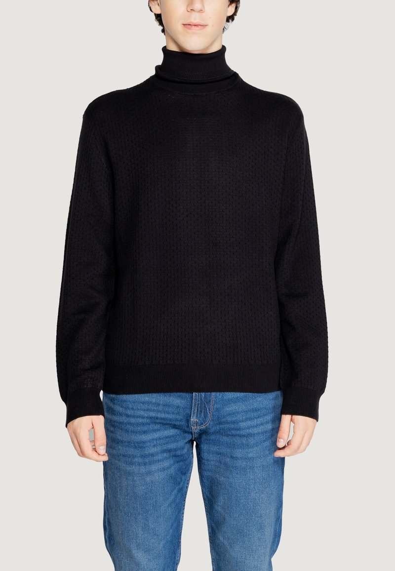 Maglione a collo alto nero con un design a maglia strutturata, caratterizzato da maniche lunghe e polsini a costine, abbinato a jeans blu.