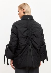 Giacca a vento nera con colletto alto, retro arricciato e maniche lunghe. Presenta nastri e una texture liscia e opaca.