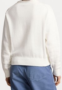 Bílý pletený svetr s žebrovaným lemem a manžetami, s jednoduchým, texturovaným vzorem. Na sobě modré kalhoty, pohled zezadu.