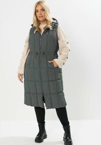 Gilet matelassé gris avec capuche, fermeture éclair à l'avant, taille cintrée à cordon, et poches latérales. Porté par-dessus un pull beige et un legging noir.