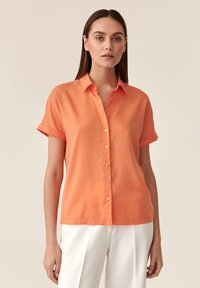 Blusa arancione a maniche corte con fronte a bottoni, tessuto morbido, maniche arrotolate e vestibilità rilassata. Abbinate a pantaloni bianchi.
